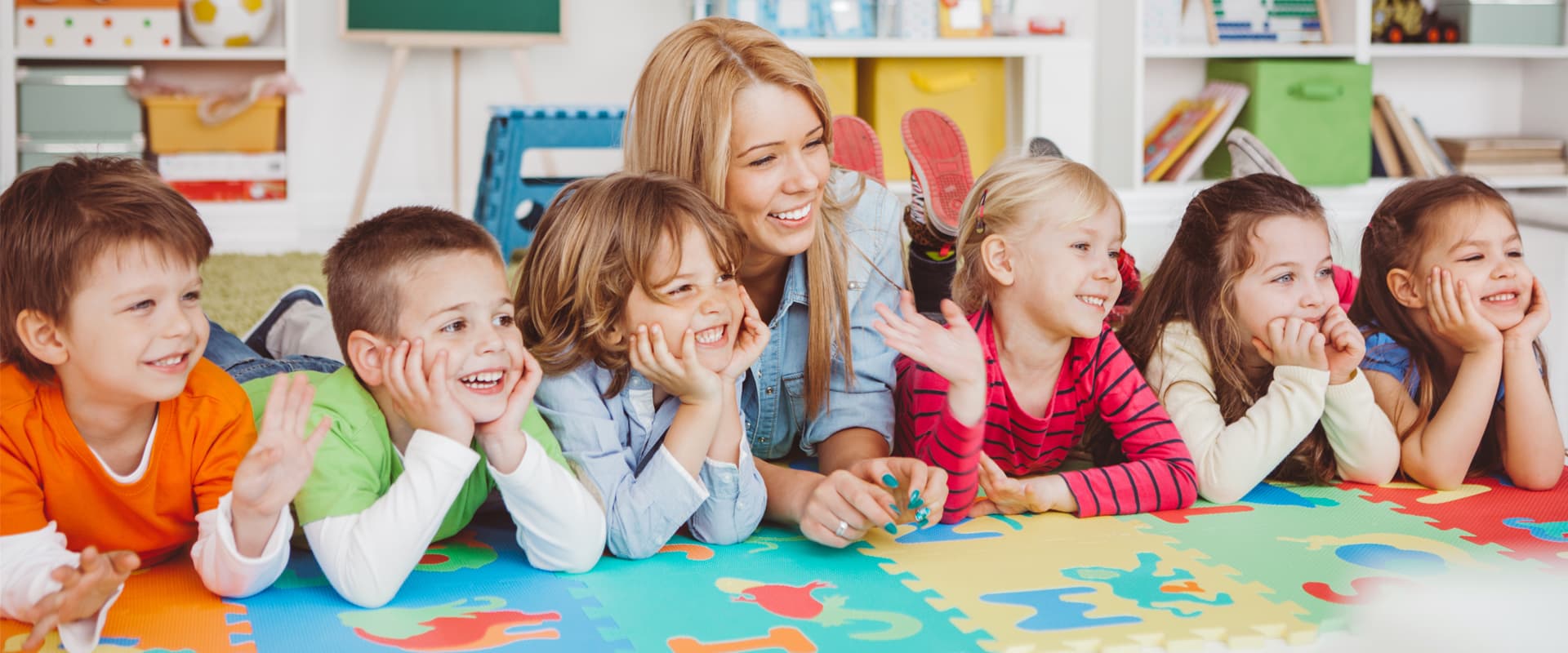 Educatoare și cinci copii zâmbind, întinși pe un covor colorat în sala de activități